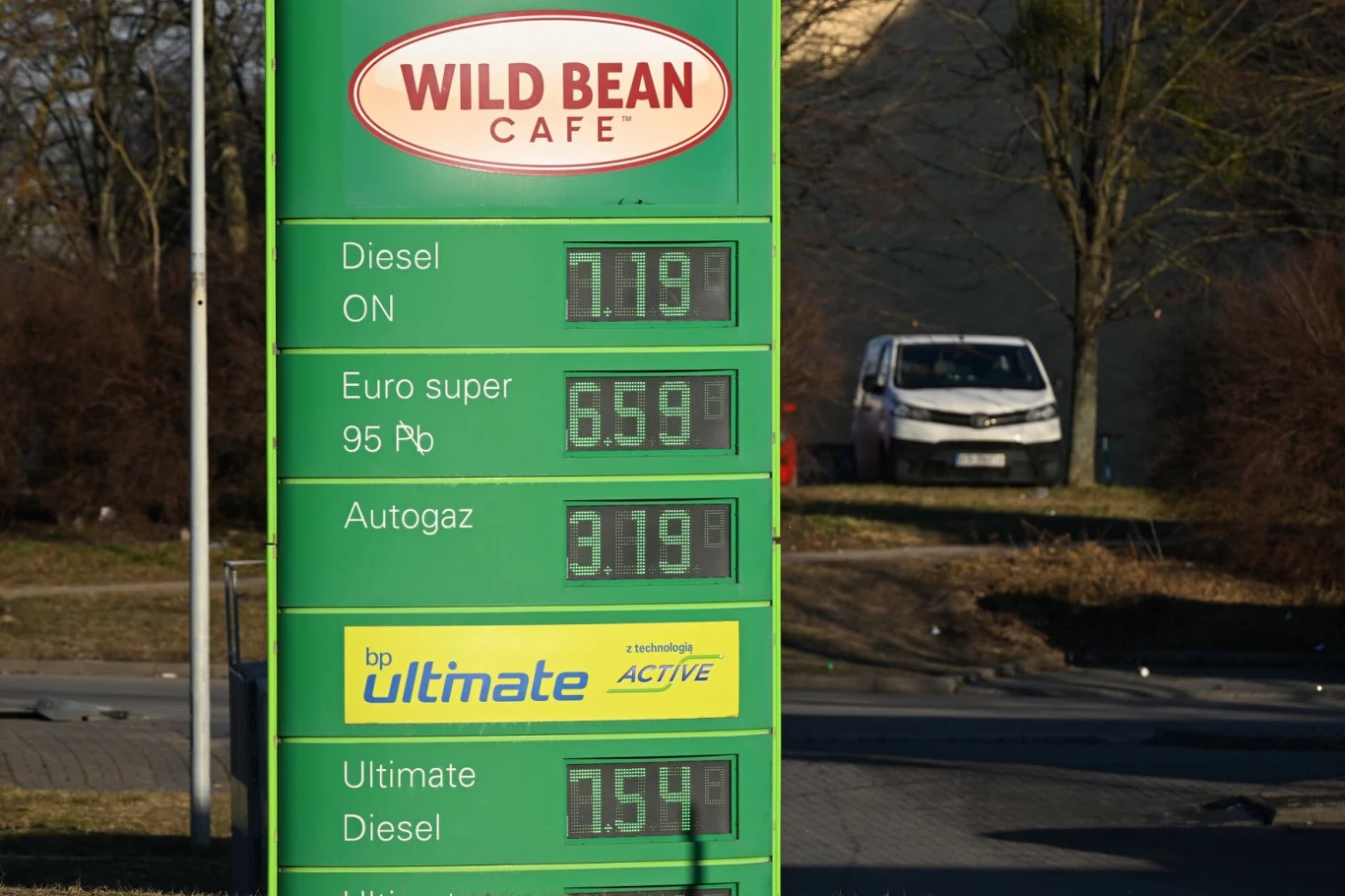 Ceny paliw na stacji BP Zielona tablica informacyjna stacji paliw z widocznymi aktualnymi cenami różnych rodzajów paliwa, w tle zaparkowany biały samochód i drzewo bez liści.