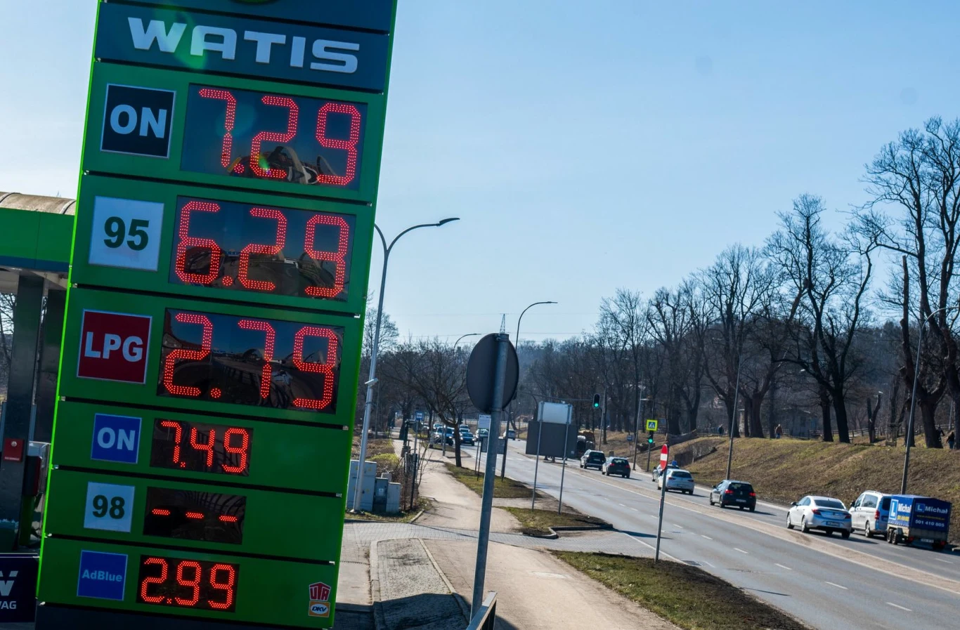 Cena paliwa na stacji Watis Tablica cenowa stacji paliw Watis z wyświetlonymi cenami różnych rodzajów paliwa, w tle droga z poruszającymi się samochodami oraz drzewami bez liści.