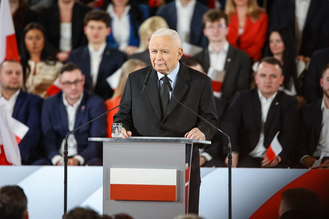 Starszy mężczyzna w garniturze przemawia przy mównicy ozdobionej polską flagą, na tle licznej grupy osób trzymających biało-czerwone flagi.