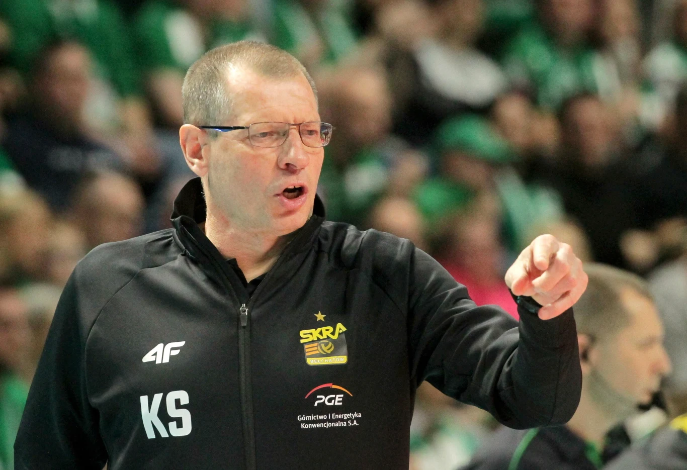 Trener drużyny siatkarskiej Skra Bełchatów w czarnej kurtce z inicjałami KS, gestykulujący i wydający polecenia podczas meczu, w tle rozmyta publiczność w zielonych barwach.
