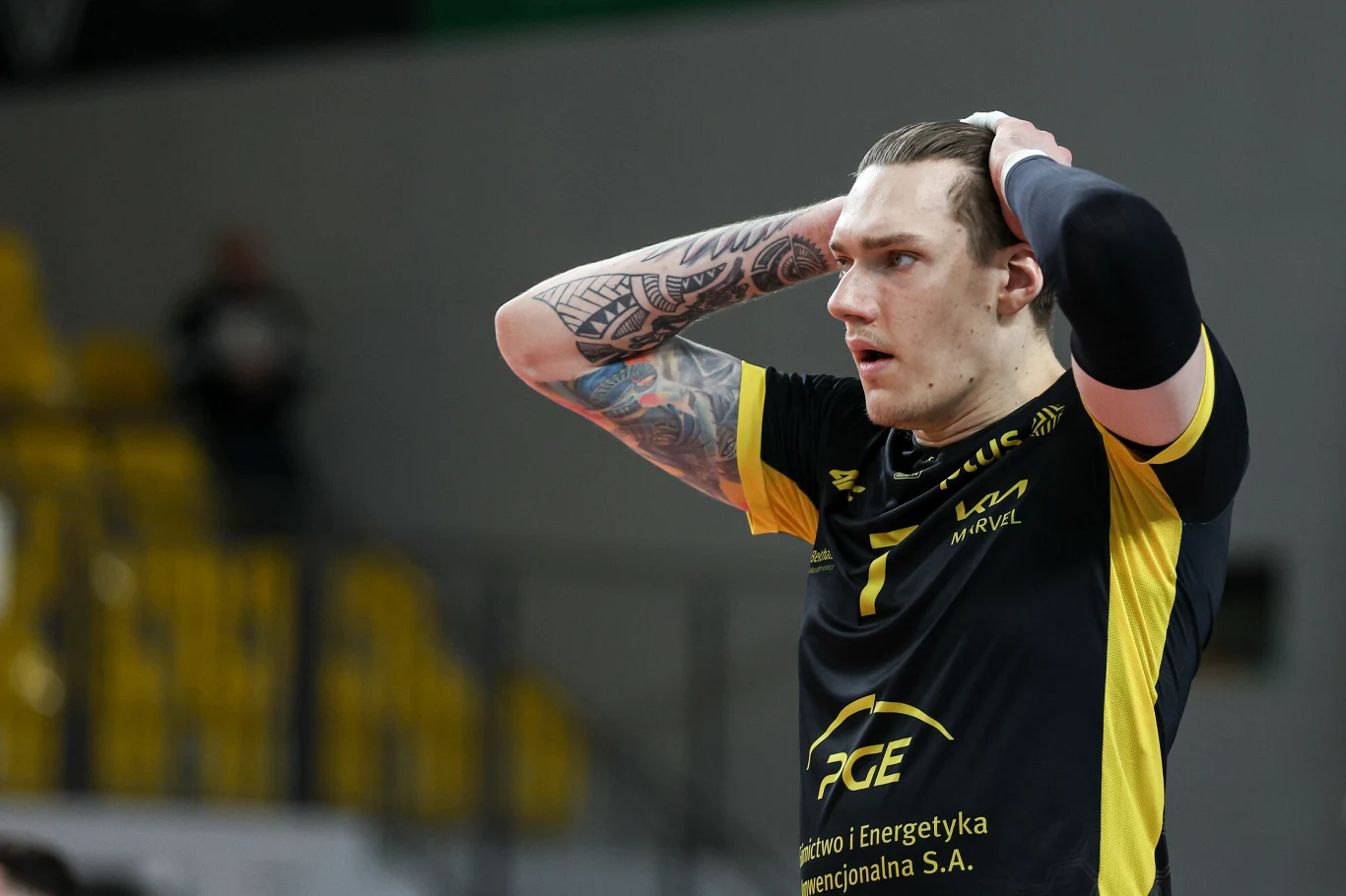 Siatkarz w żółto-czarnym stroju z tatuażami na rękach trzyma dłonie na głowie w wyrazie zaskoczenia lub zdenerwowania, w tle rozmyte żółte krzesełka trybun.