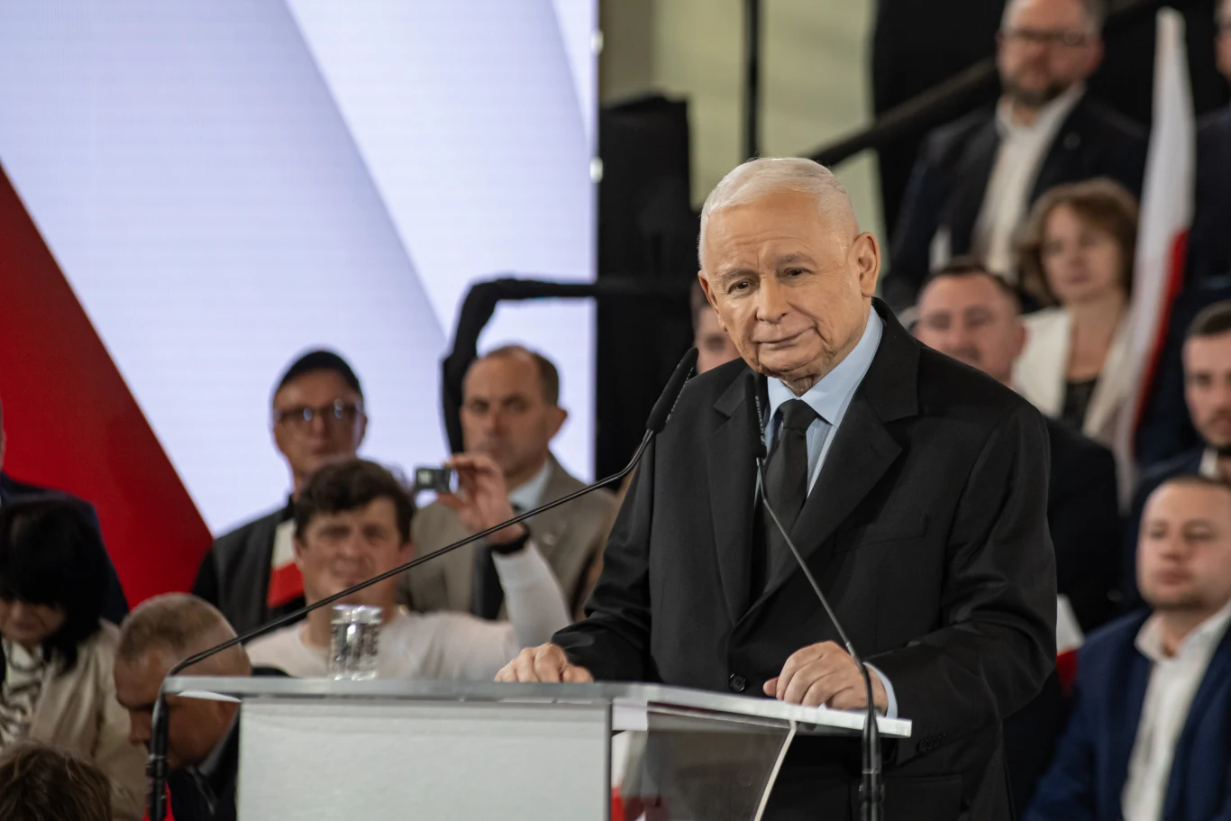 Starszy mężczyzna w czarnym garniturze przemawia przy przezroczystej mównicy na scenie, otoczony grupą słuchających go osób na tle biało-czerwonych elementów.