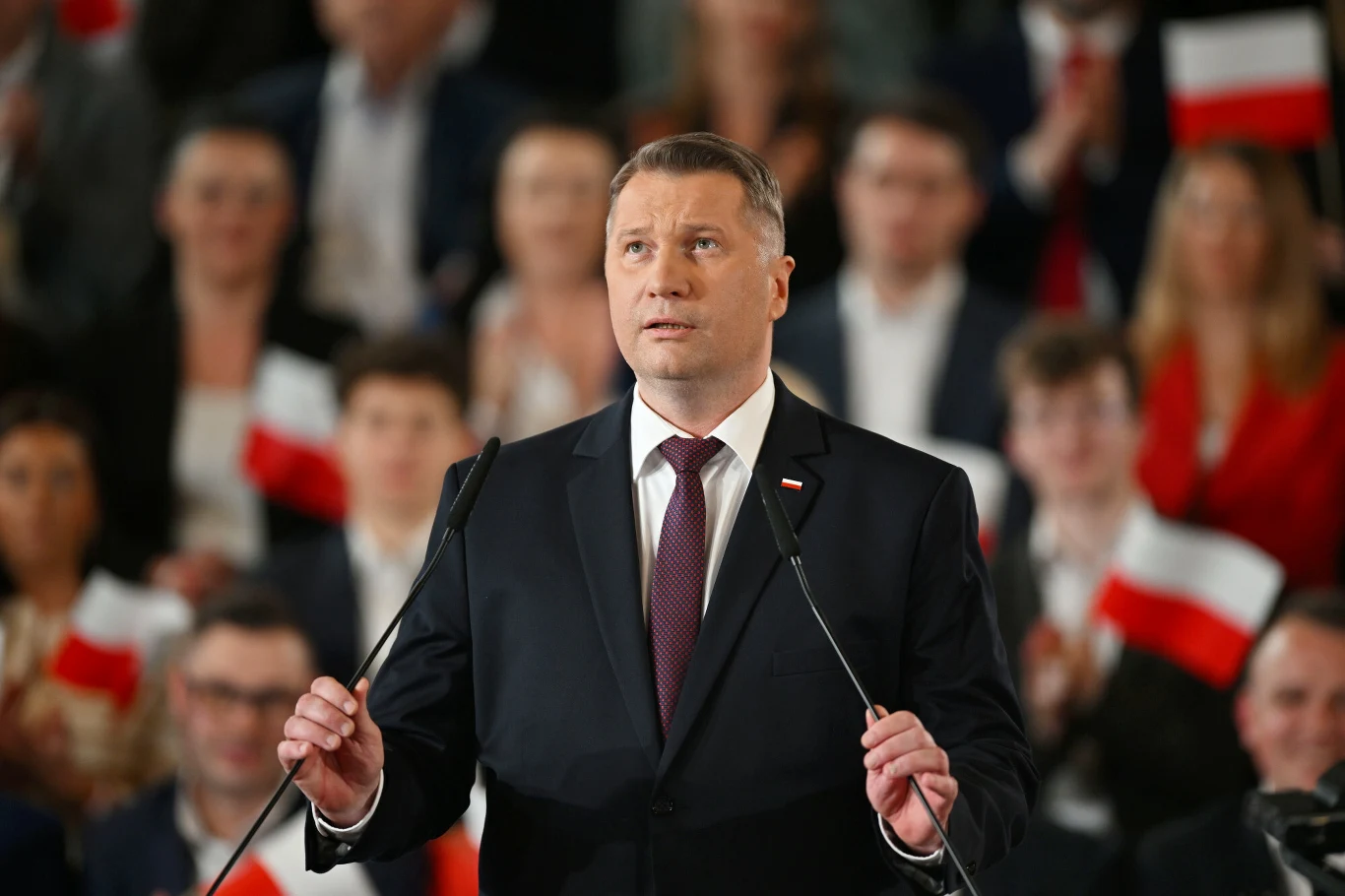 Przemysław Czarnek podczas konwencji PiS w Krakowie Mężczyzna w garniturze i bordowym krawacie stoi przy mównicy, trzymając mikrofony, na tle tłumu ludzi z polskimi flagami w rękach.