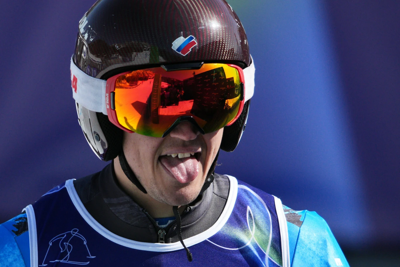 Narciarz w kasku i sportowych okularach z przyciemnianymi szkłami, wystawiający język, ubrany w strój zawodniczy z numerem startowym.