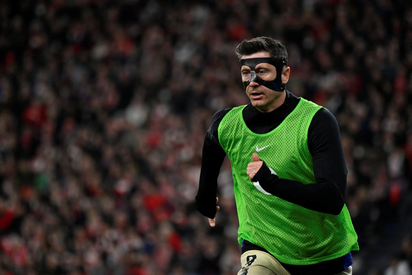 Piłkarz w zielonej kamizelce treningowej biegnie po murawie, mając na twarzy ochronną czarną maskę, w tle rozmyta publiczność stadionu.