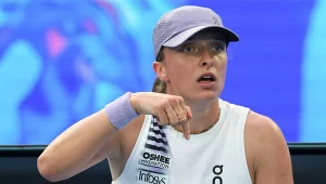 Tenisistka w białej koszulce bez rękawów i fioletowej czapce z daszkiem trzyma ręcznik i wskazuje palcem w dół, wydaje się być zaangażowana w rozmowę; w tle rozmazane, niebieskie elementy kortu tenisowego.