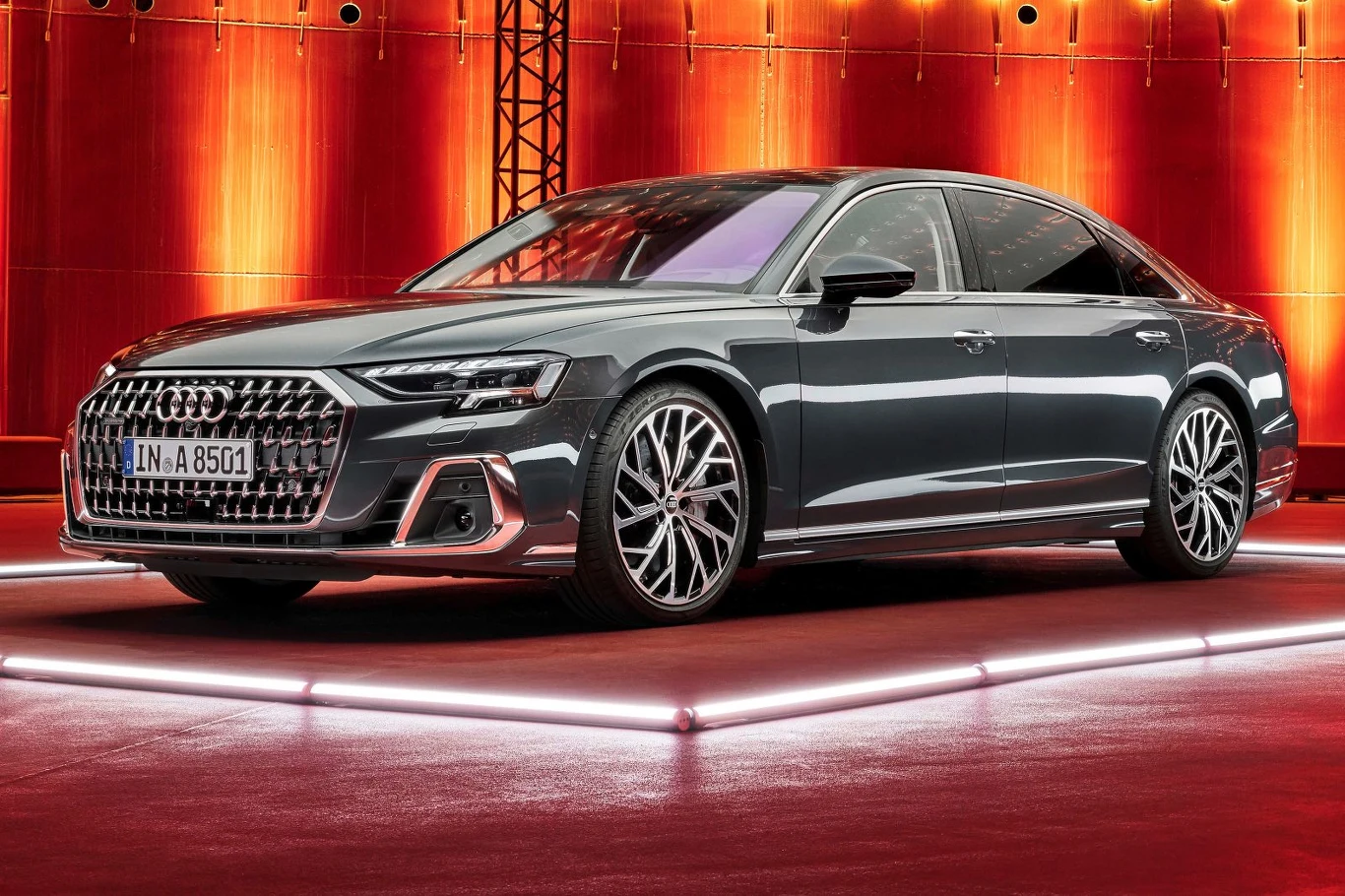 Luksusowy sedan Audi A8 w kolorze grafitowym, ustawiony na podświetlonej platformie, z charakterystyczną dużą osłoną chłodnicy i eleganckimi, wieloramiennymi felgami, na tle nowoczesnej, czerwonej scenografii.