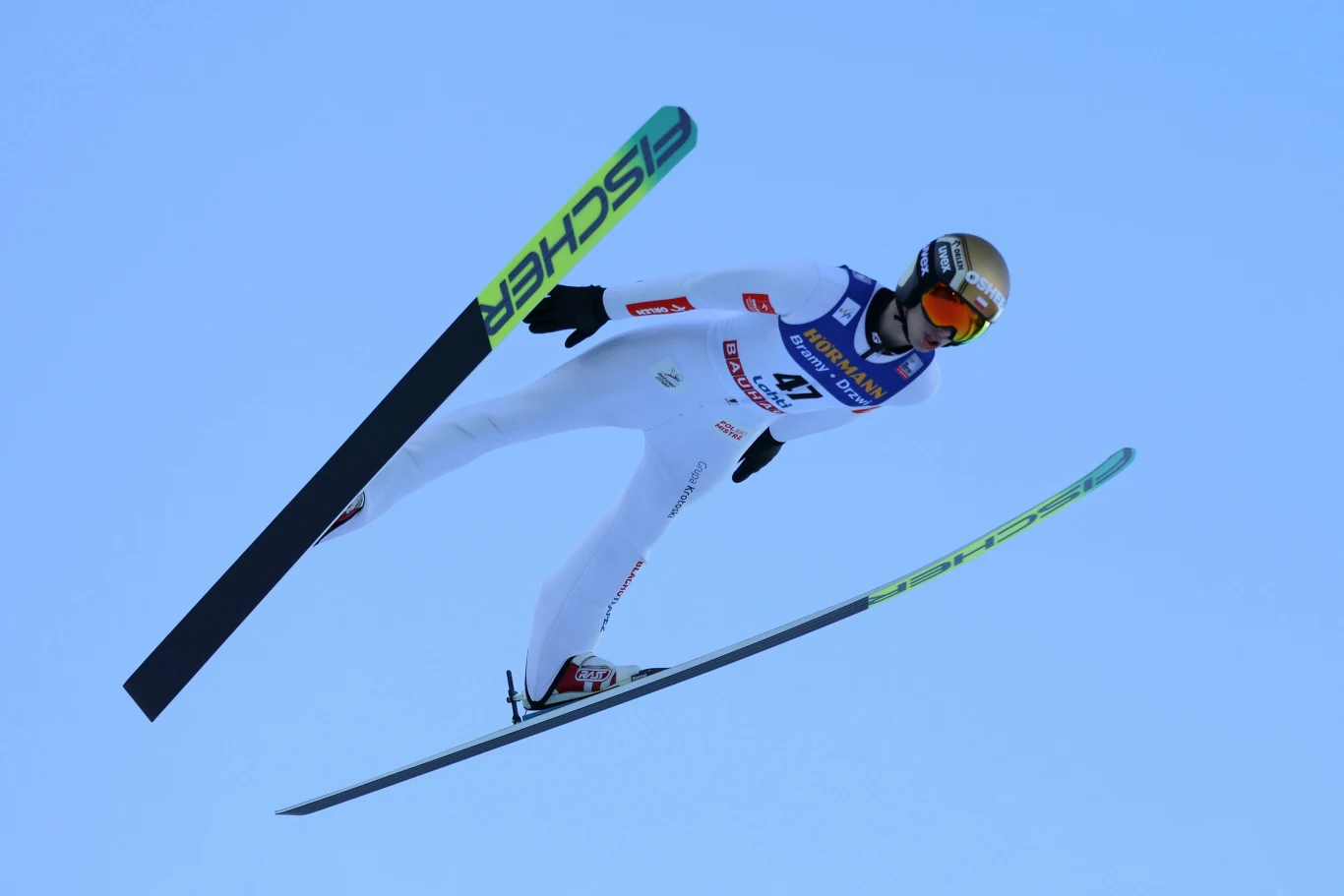 Kacper Tomasiak w locie na nartach, biały kombinezon, złoty kask, gogle, szeroko rozstawione narty w V.