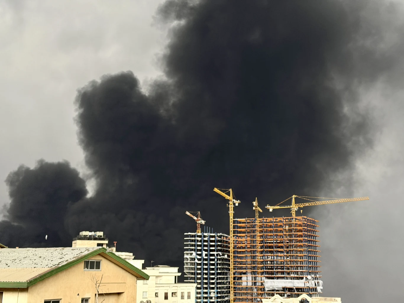 Gęsty czarny dym unoszący się nad placem budowy z wysokimi dźwigami i konstrukcjami budynków na tle pochmurnego nieba.