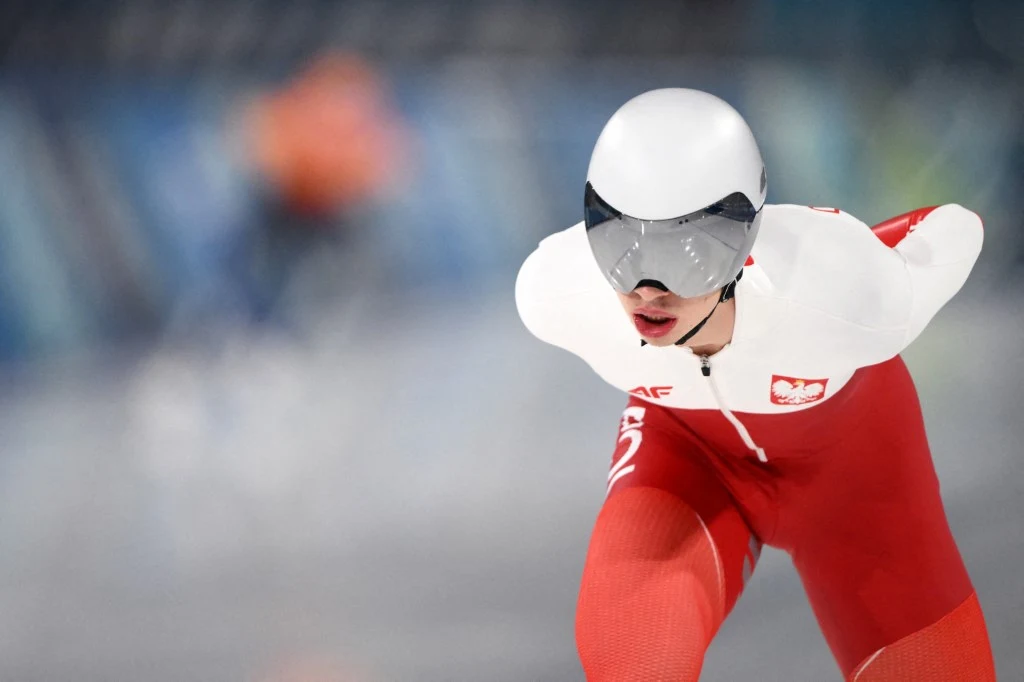 Łyżwiarz szybki w biało-czerwonym kombinezonie z orzełkiem na piersi, w trakcie wyścigu na lodowisku, w aerodynamicznej pozycji z pochyloną sylwetką i rękami za plecami.