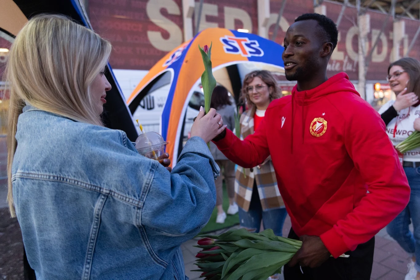 Mężczyzna w czerwonej bluzie wręcza kobiecie tulipana, obok stoją uśmiechnięte osoby, a w tle widoczny jest namiot z logotypem STS.