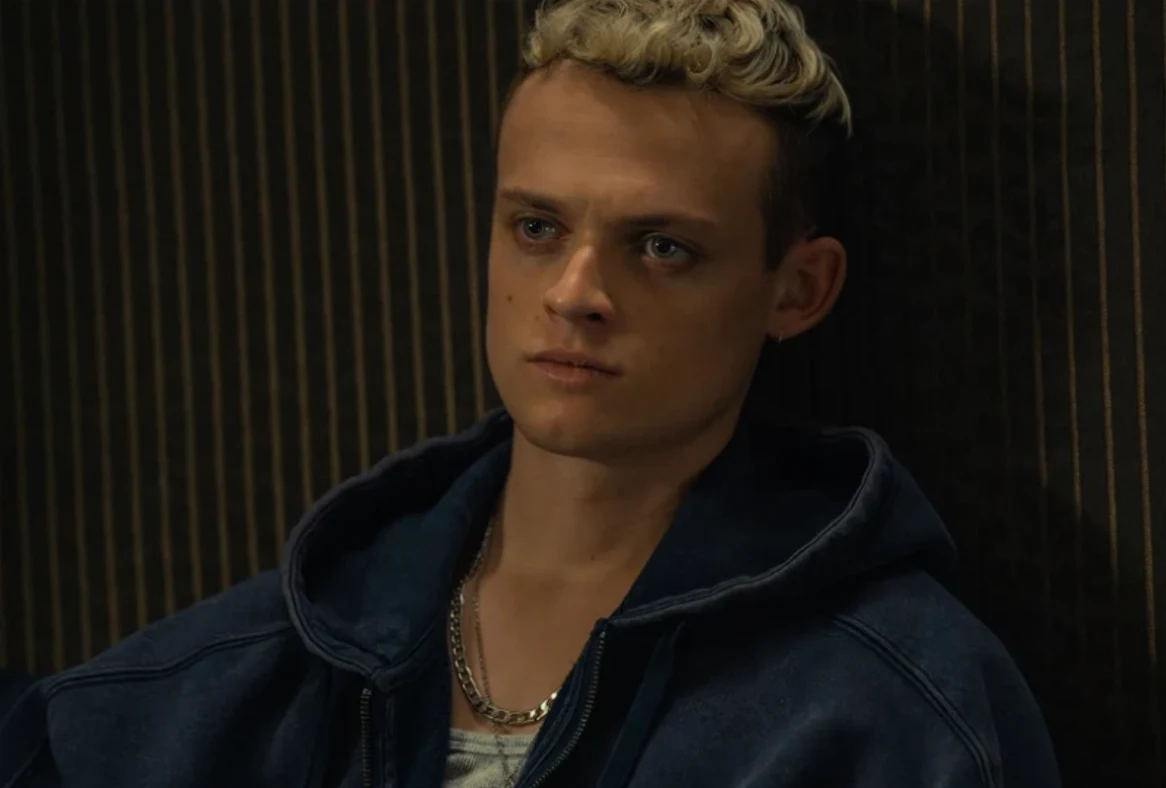 Młody mężczyzna o krótkich, kręconych blond włosach i poważnym wyrazie twarzy siedzi w ciemnoniebieskiej bluzie z kapturem, w tle widoczne pionowe prążkowane linie.