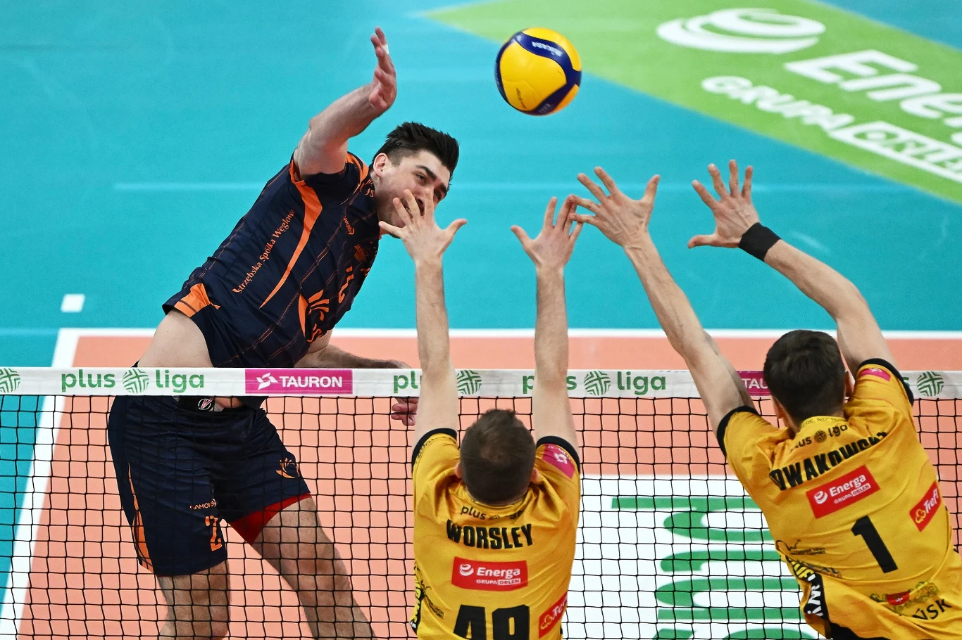 Siatkarz w granatowo-pomarańczowym stroju atakuje piłkę nad siatką, podczas gdy dwóch zawodników w żółtych koszulkach próbuje zablokować uderzenie, unosząc ręce do góry tuż przy siatce na tle hali sportowej z widocznymi reklamami na boisku.