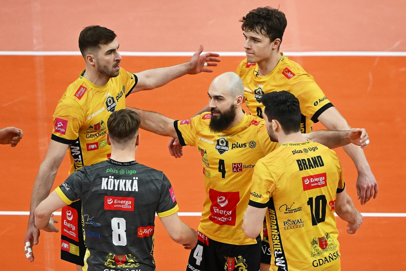 Grupa siatkarzy w żółtych i czarnych koszulkach celebruje punkt na boisku do siatkówki, skupiając się na wzajemnym wsparciu i energii zespołu.