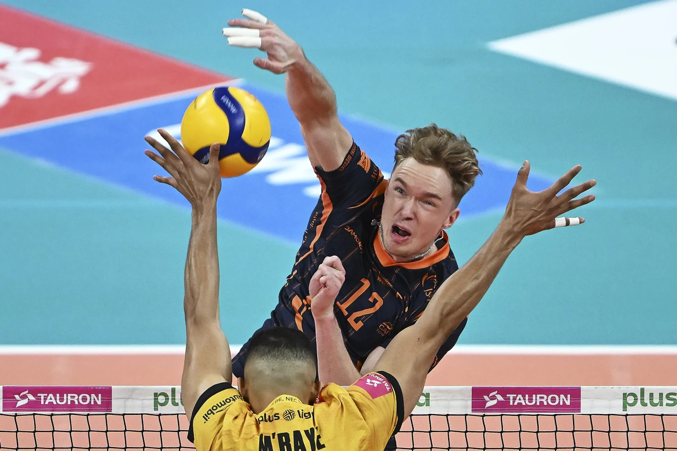 Siatkarz w czarno-pomarańczowym stroju dynamicznie atakuje piłkę nad siatką, podczas gdy zawodnik w żółtej koszulce próbuje go zablokować, obaj skupieni na grze w hali sportowej.