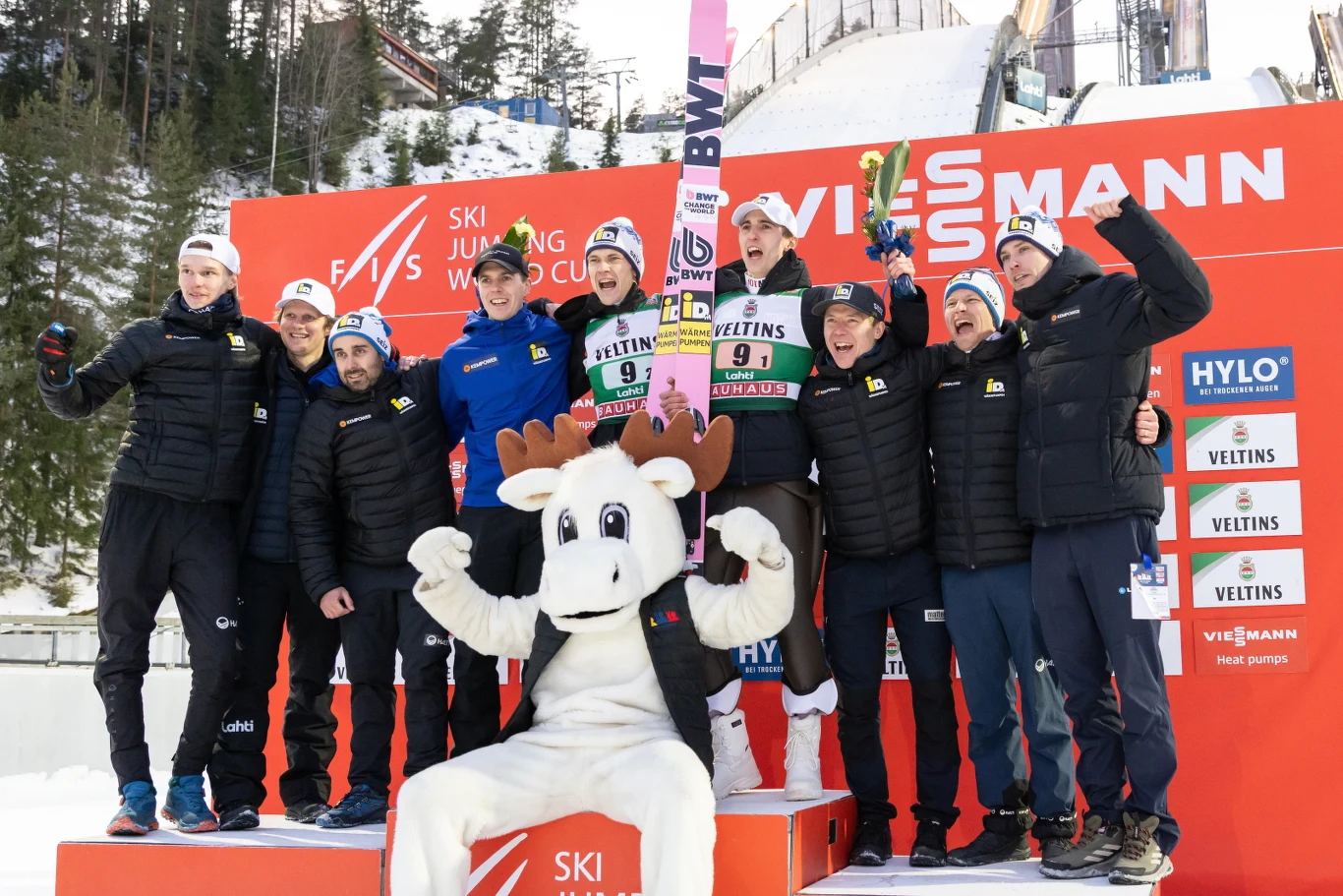 Grupa skoczków narciarskich oraz członków sztabu drużyny pozuje razem na podium zwycięzców zawodów Pucharu Świata, obok nich maskotka w stroju łosia, w tle widoczna duża skocznia narciarska oraz logotypy sponsorów na czerwonym tle.