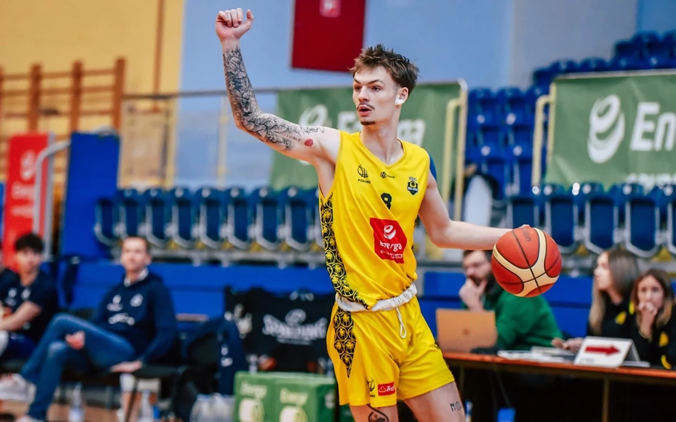 Koszykarz w żółtym stroju trzyma piłkę jedną ręką i unosi drugą w górę, sygnalizując zagranie podczas meczu na hali sportowej.