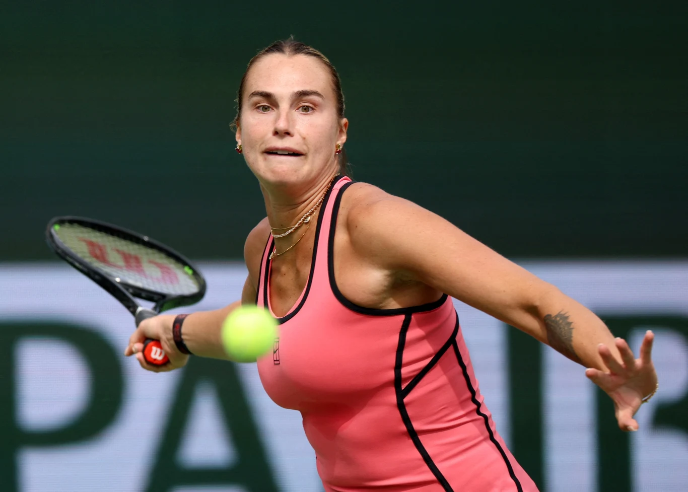 Kobieta w sportowym stroju tenisowym w trakcie dynamicznego uderzenia piłki rakietą, skoncentrowana twarz i widoczny ruch ramienia, tło rozmyte z dominującym ciemnozielonym kolorem.