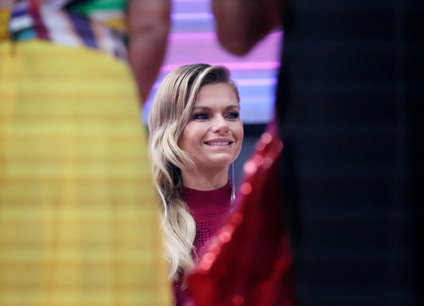 Uśmiechnięta kobieta z długimi, jasnymi włosami w bordowej sukience, otoczona sylwetkami innych osób w kolorowych ubraniach, z rozmytym, fioletowym tłem.