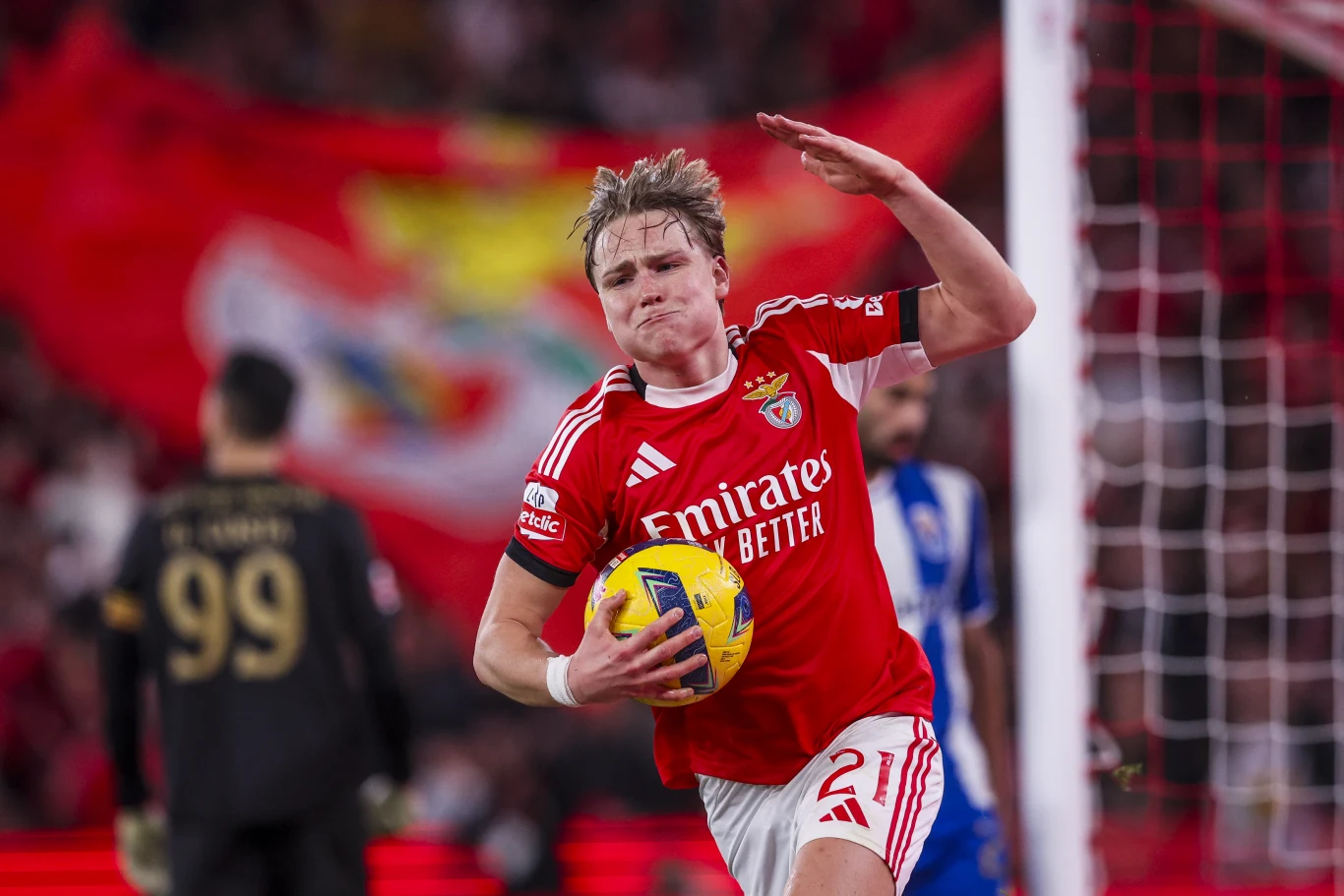 Piłkarz drużyny Benfica w czerwonym stroju ściska piłkę pod pachą i unosi rękę, za nim widoczny bramkarz w czarnym stroju oraz duża, rozmazana flaga klubu w tle.