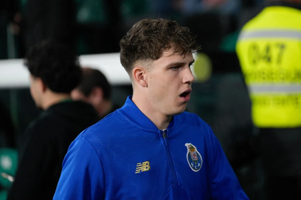 Młody mężczyzna w niebieskiej sportowej kurtce z herbem klubu piłkarskiego FC Porto stoi na stadionie, patrząc w bok; w tle widoczna jest postać w żółtej kamizelce odblaskowej.