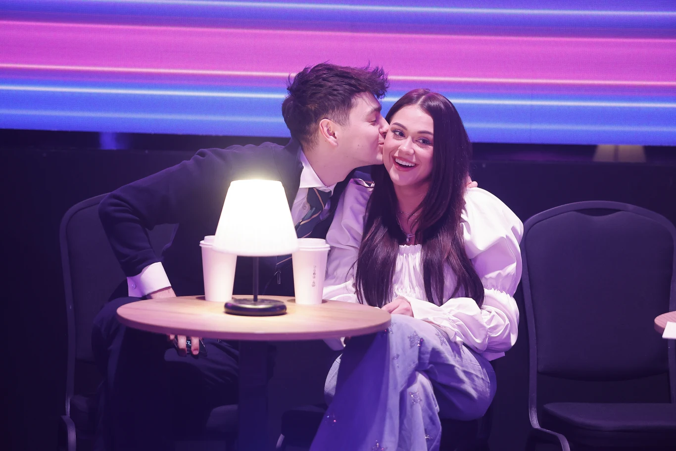 Para siedząca przy małym okrągłym stoliku, mężczyzna całuje kobietę w policzek, a ona szeroko się uśmiecha; na stoliku stoją dwie papierowe kubki i zapalona lampka, w tle wyraźne, kolorowe światła.