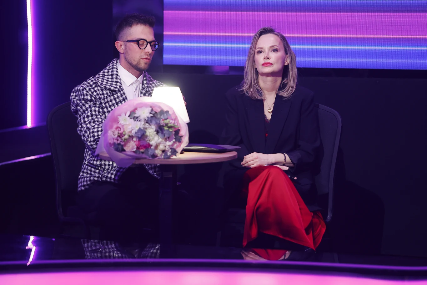 Dwie osoby siedzą przy okrągłym stoliku, na którym znajduje się bukiet kwiatów i jasne światło, otoczeni fioletowym oświetleniem; kobieta ubrana w czarny żakiet i czerwone spodnie patrzy poważnie przed siebie, mężczyzna w okularach i kraciastej marynar...