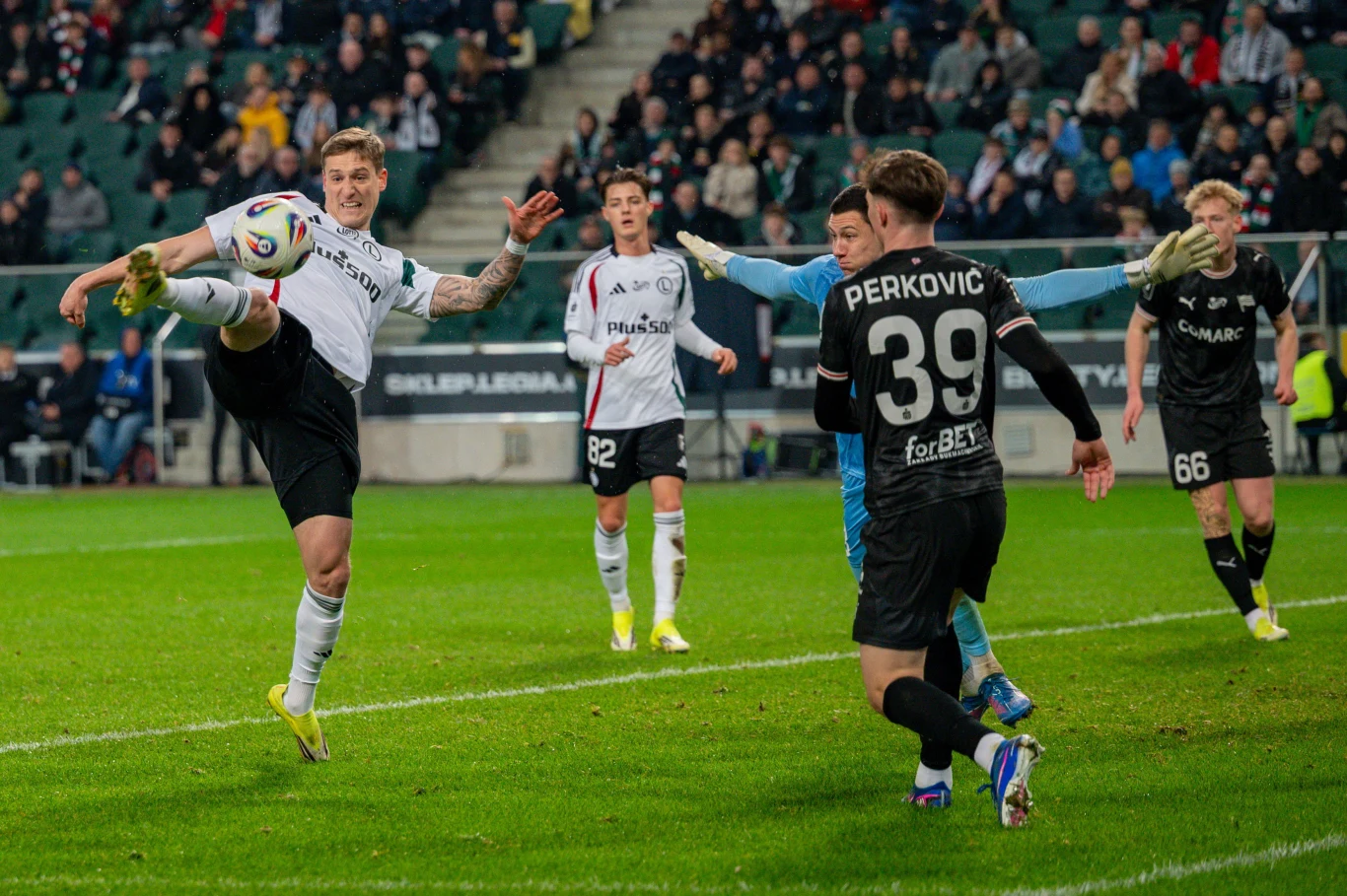 Piłkarz w białej koszulce wykonuje efektowny wykop piłki w powietrzu, wokół niego znajduje się kilku zawodników drużyny przeciwnej oraz bramkarz, a całość rozgrywa się na tle zatłoczonych trybun stadionu piłkarskiego.