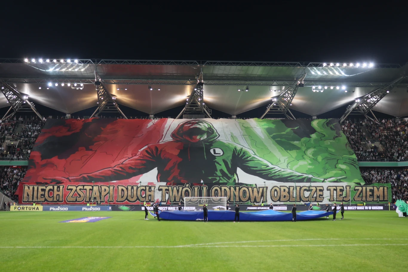 Gigantyczna sektorówka z postacią w kapturze ubranej w barwy klubowe na tle zielono-czerwonym, z dużym napisem „NIECH ZSTĄPI DUCH TWÓJ I ODNOWI OBLICZE TEJ ZIEMI” podczas meczu piłkarskiego, kibice wypełniający trybuny stadionu.