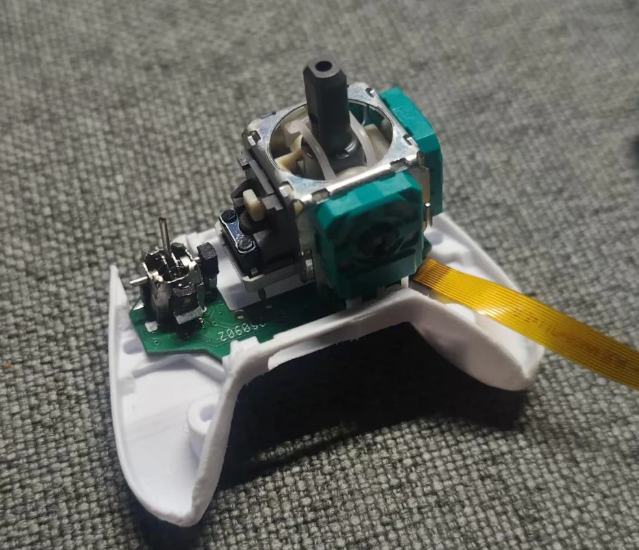 Moduł analogowego drążka kontrolera z zamontowaną płytką PCB, zielonym potencjometrem i podłączoną taśmą fleksyjną, umieszczony na białej, częściowo zdemontowanej obudowie, całość leży na szarej tkaninie.