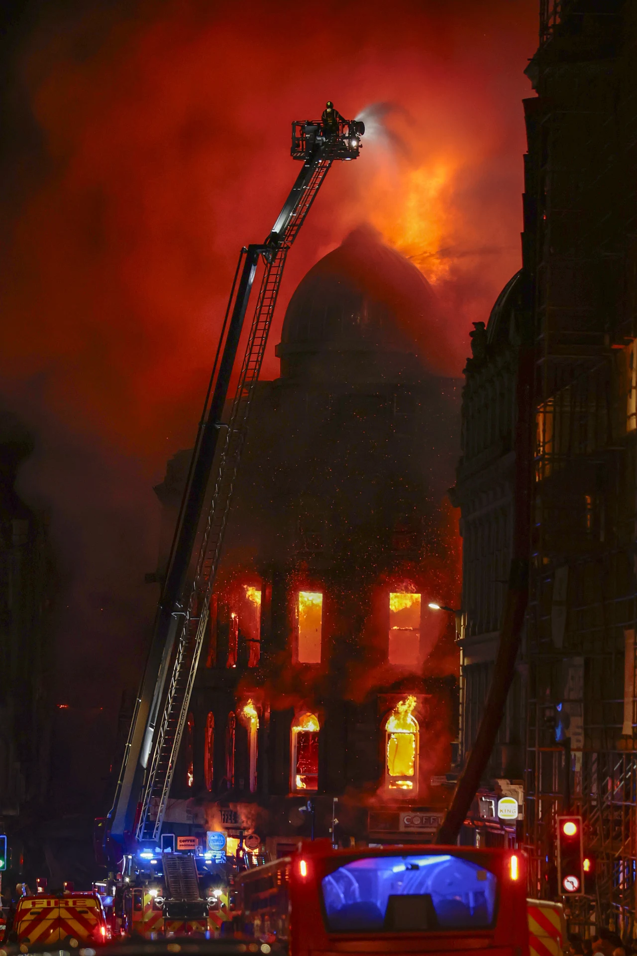 Intensywny pożar trawiący wielokondygnacyjny budynek, płomienie wydostają się przez okna i dach, strażacy na wysięgniku walczą z ogniem, ulica oświetlona światłami pojazdów ratowniczych.