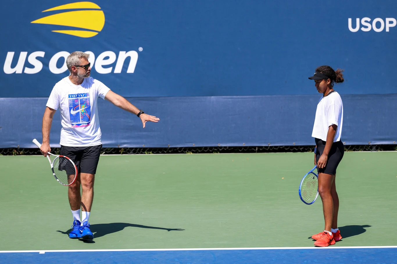 Mężczyzna wykonujący gest ręką instruuje młodą kobietę podczas treningu tenisowego na korcie US Open, oboje trzymają rakiety tenisowe i noszą sportowe ubrania.