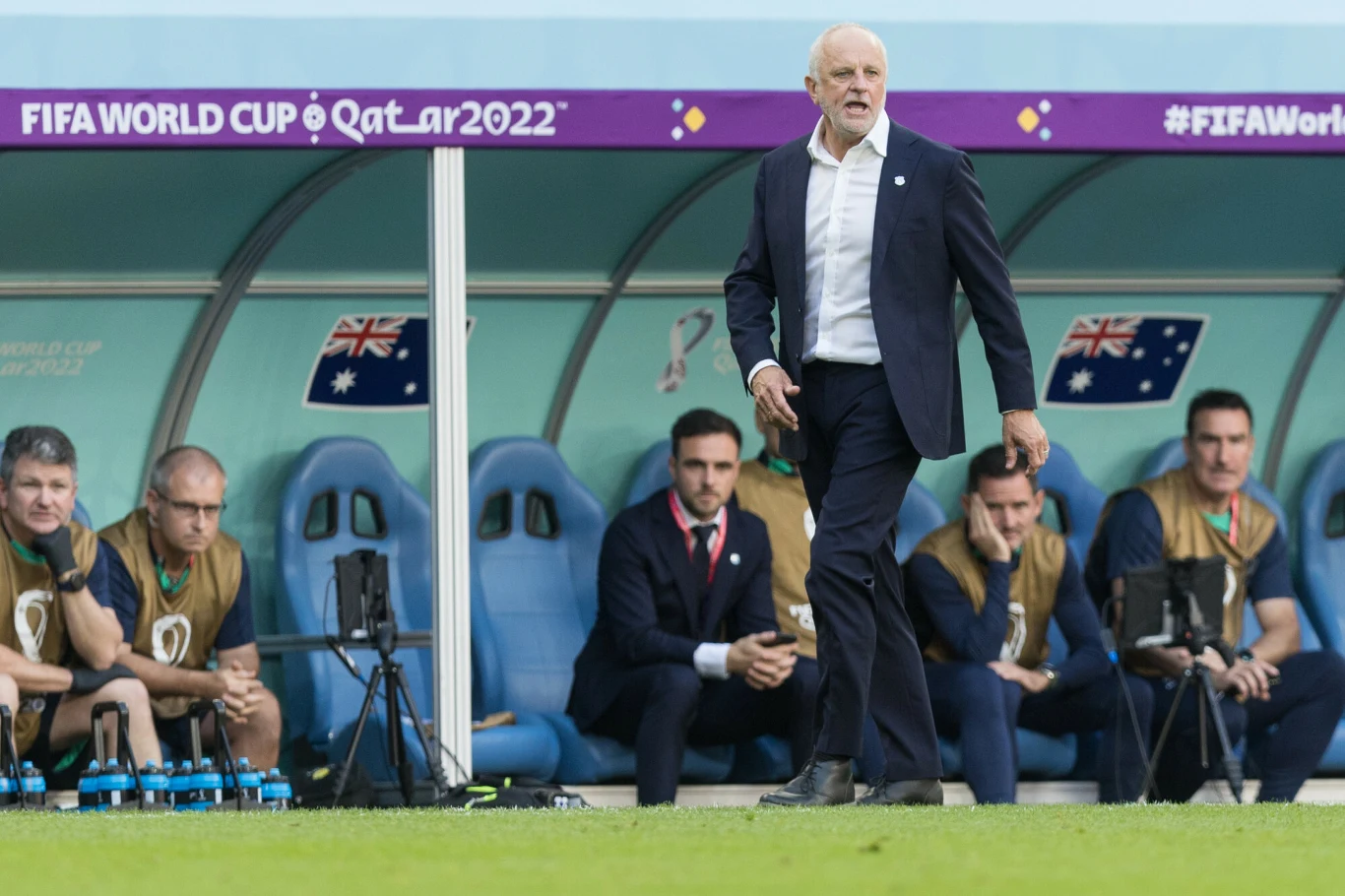 Trener w ciemnym garniturze energicznie gestykuluje przy linii bocznej boiska piłkarskiego, za nim na ławce rezerwowych siedzą członkowie sztabu szkoleniowego oraz piłkarze reprezentacji Australii, a w tle widoczne są flagi i napisy związane z Mistrzos...