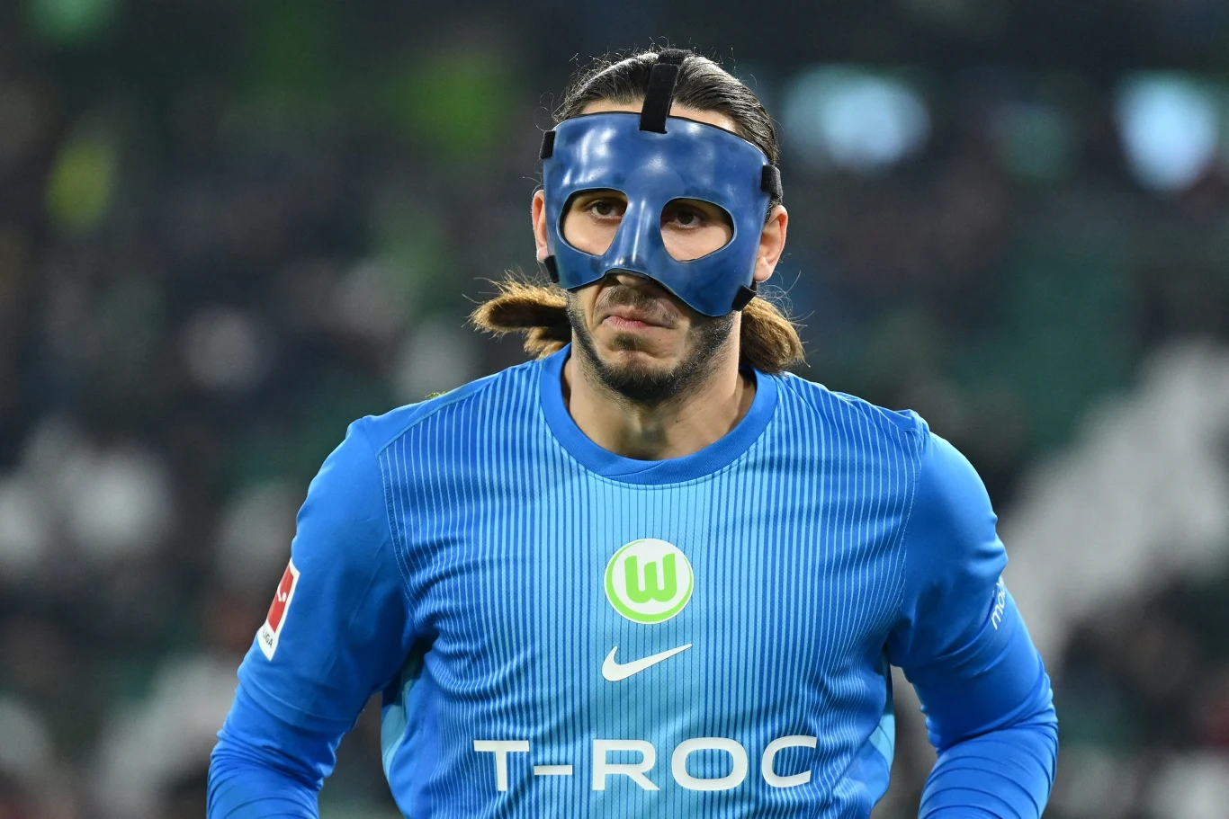 Piłkarz w niebieskiej koszulce Wolfsburga z logotypami sponsora i klubu, mający na twarzy czarną ochronną maskę, stoi na tle nieostrego stadionu podczas meczu.
