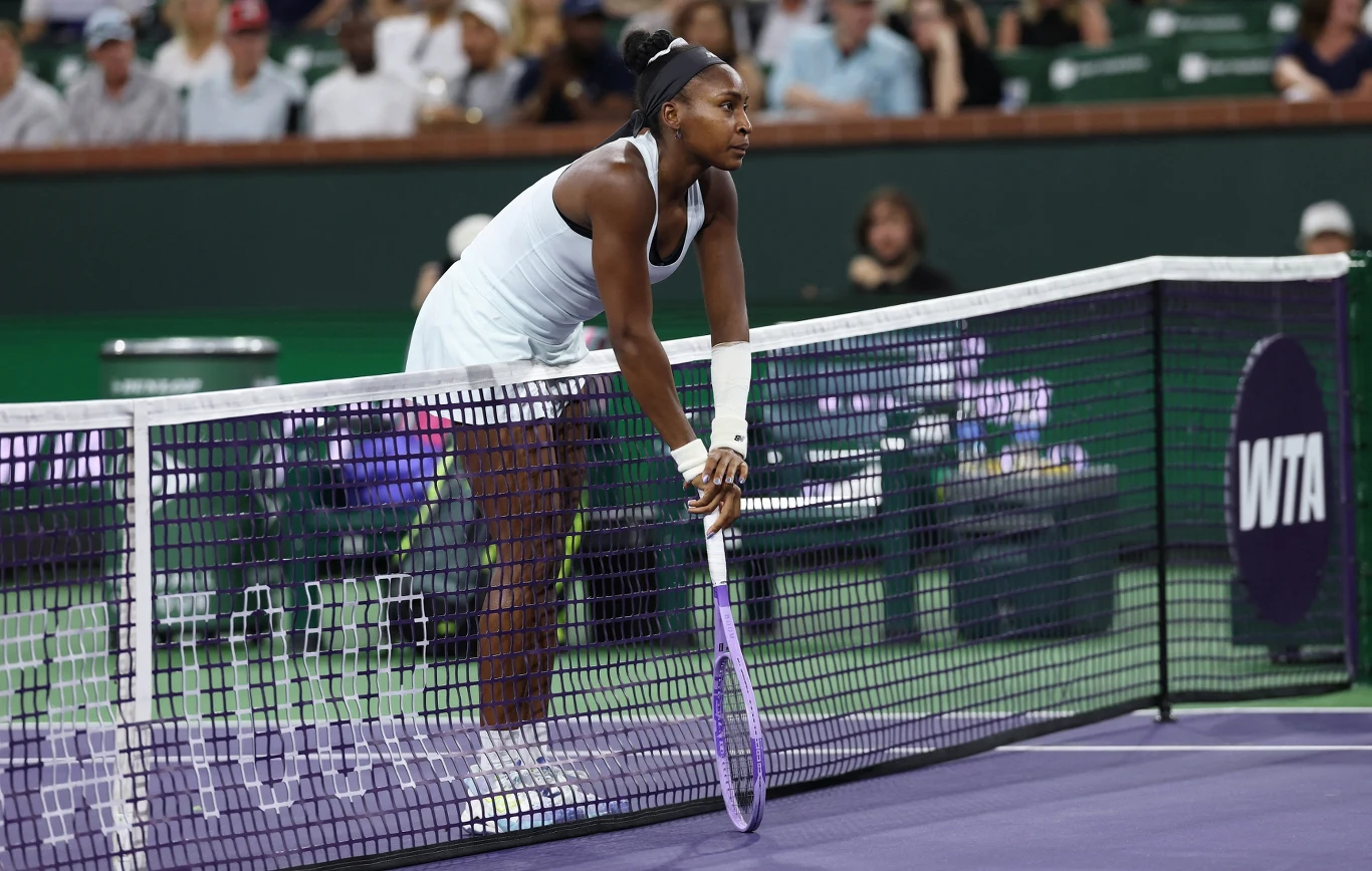 Tenisistka w białym stroju oparta o siatkę kortu, trzymająca rakietę, skupiona na grze, na tle publiczności i logotypu WTA.