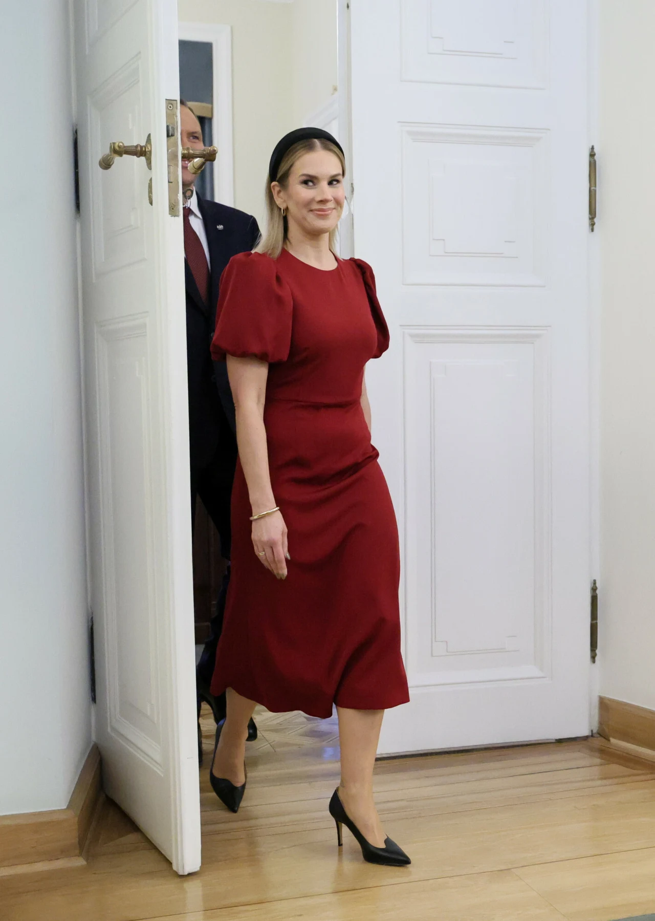 Kobieta w eleganckiej, bordowej sukience i czarnych szpilkach wchodzi do jasnego pomieszczenia przez otwarte białe drzwi, za nią znajduje się mężczyzna w garniturze.