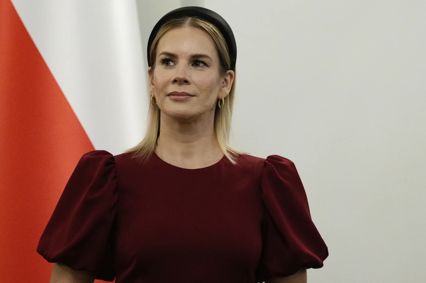 Kobieta w bordowej sukience z bufiastymi rękawami i czarną opaską na włosach stoi na tle fragmentu biało-czerwonej flagi.