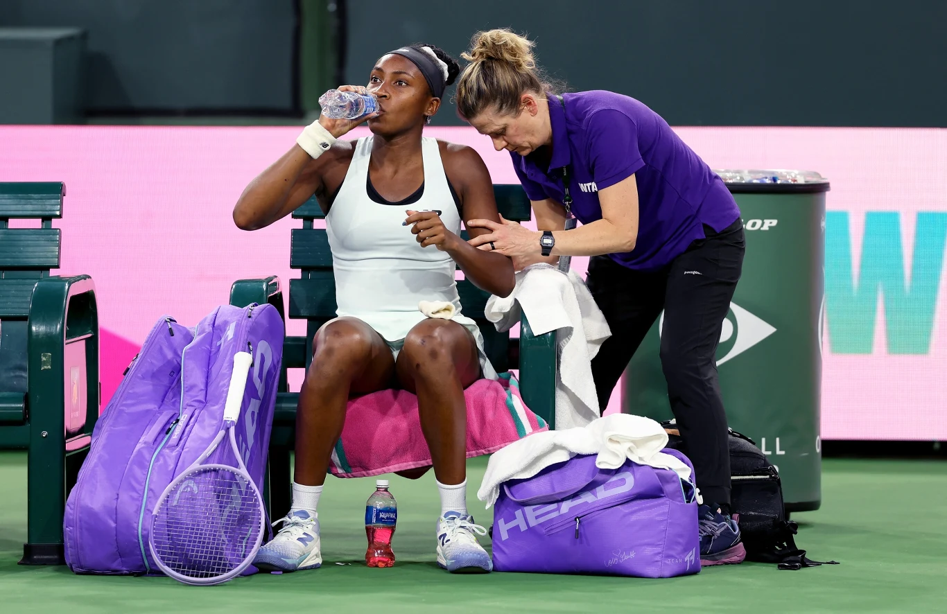 Tenisistka siedzi na ławce z butelką wody w ręku, obok niej leży rakieta tenisowa i torby sportowe w fioletowych kolorach, a trenerka ubraną w fioletową koszulkę udziela jej wskazówek lub sprawdza notatki.
