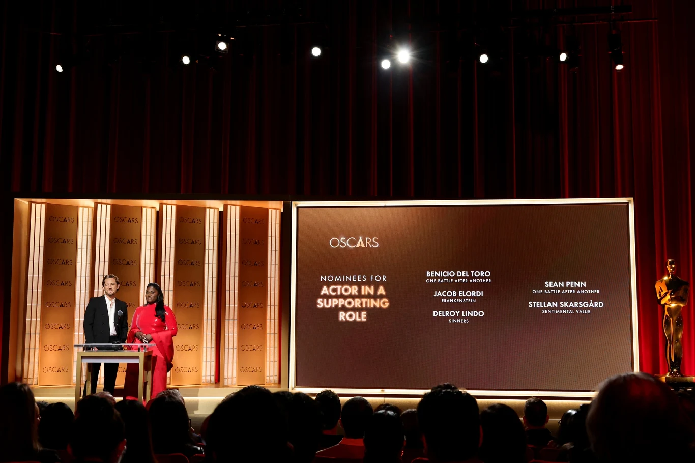 Dwóch prowadzących stojących na scenie obok dużego ekranu wyświetlającego nominacje do Oscara w kategorii Aktor Drugoplanowy, na widowni widoczne sylwetki widzów, a z boku scena ozdobiona statuetką Oscara.