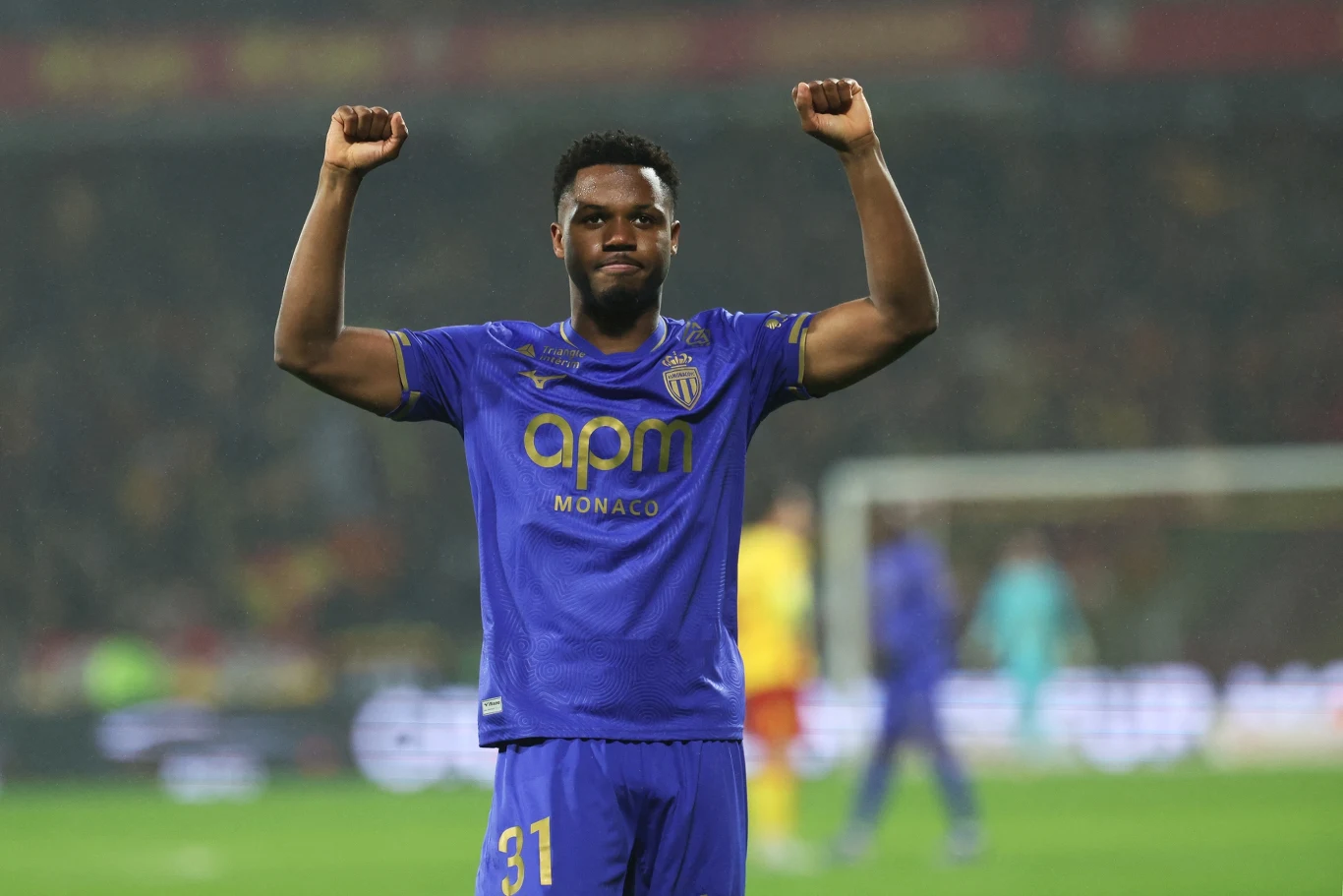 Piłkarz w fioletowej koszulce z numerem 31 i logotypem APM Monaco unosi obie ręce w geście triumfu, stojąc na boisku podczas meczu, w tle widoczni pozostali zawodnicy oraz rozmyte postaci na stadionie.