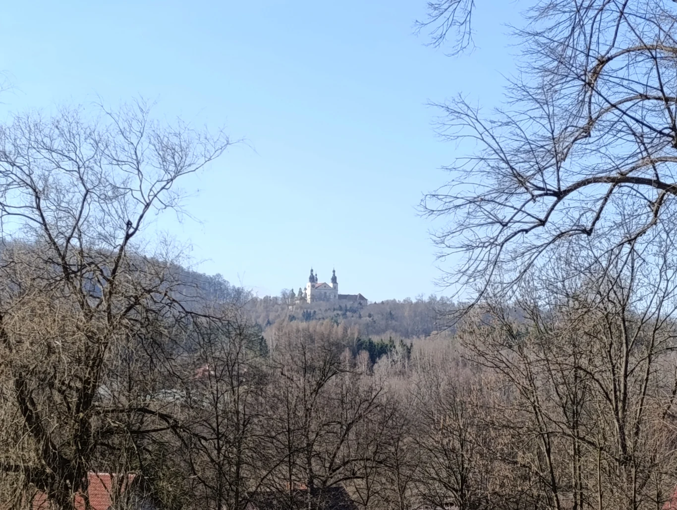 Klasztor położony na wzgórzu, otoczony drzewami bez liści, na tle jasnego, niebieskiego nieba.