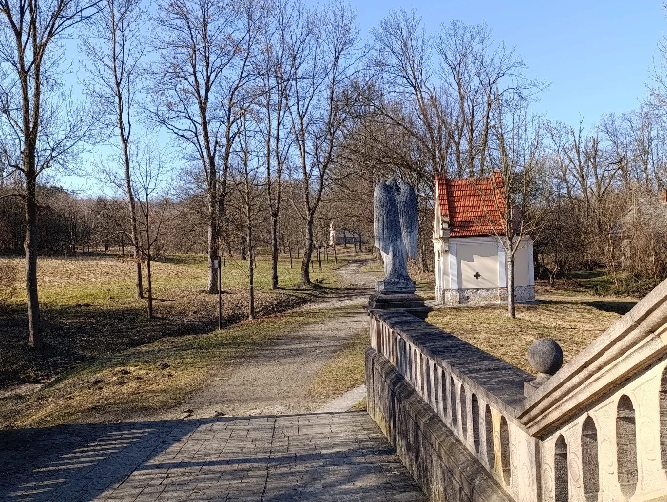 Kamienna figura anioła stojąca na końcu mostka, obok niewielka kapliczka z czerwonym dachem, otoczona bezlistnymi drzewami i ścieżką prowadzącą w głąb parku.