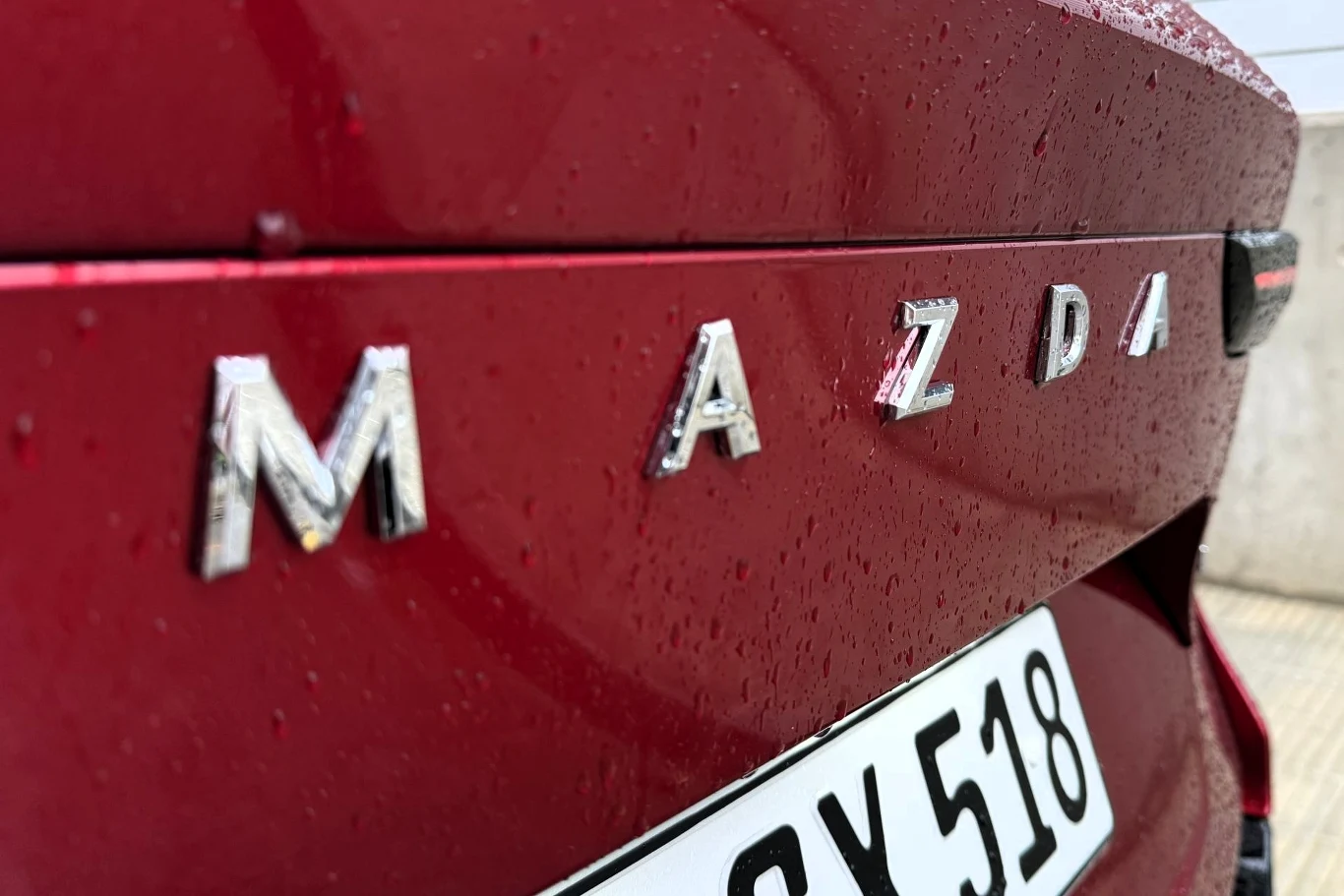 Czerwony tył samochodu marki Mazda z widocznymi chromowanymi literami logotypu oraz fragmentem tablicy rejestracyjnej, pokryty kroplami deszczu.