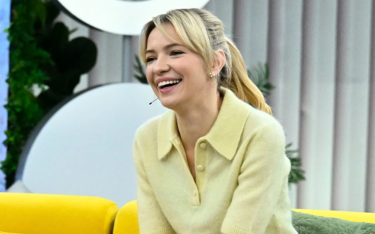 Uśmiechnięta kobieta o blond włosach spiętych w kucyk, ubrana w jasny sweter z kołnierzem, siedzi na żółtej kanapie w nowoczesnym, jasnym wnętrzu.