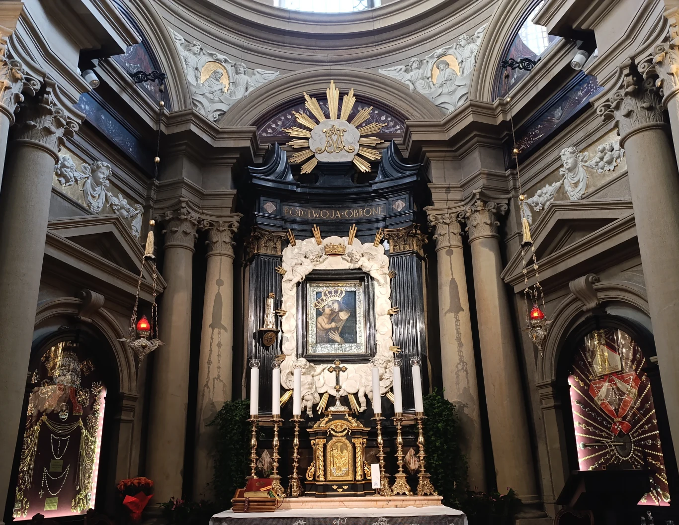 Barokowy ołtarz z obrazem Madonny z Dzieciątkiem, otoczony dekoracyjną architekturą z kolumnami, złotymi detalami i rzeźbami, podkreślony światłem wpadającym przez kopułę.