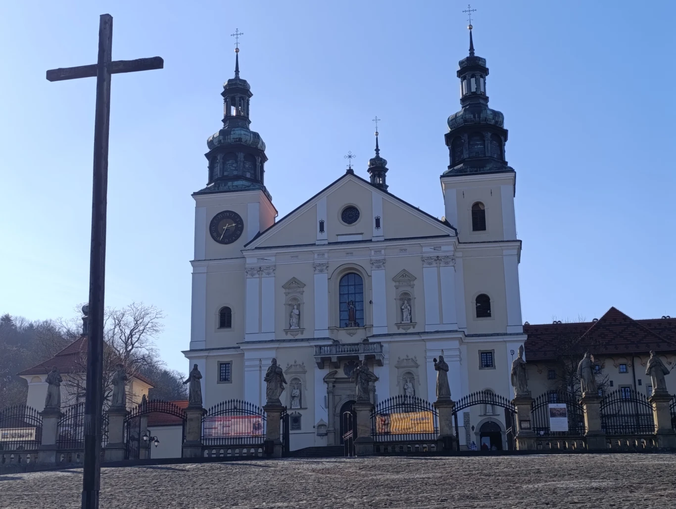 Barokowa fasada kościoła z dwoma wysokimi wieżami, bogatą dekoracją rzeźbiarską i dużym, żelaznym krzyżem w centralnej części pierwszego planu; otoczenie uzupełniają ozdobne ogrodzenia i fragmenty zabudowań klasztornych.