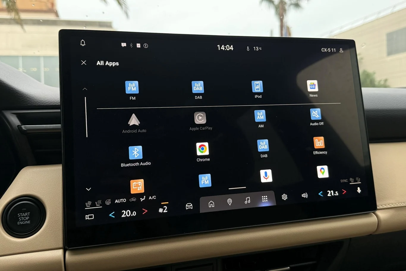 Duży ekran dotykowy systemu multimedialnego samochodu z wyraźnie widocznymi ikonami aplikacji takich jak FM, DAB, Android Auto, Apple CarPlay, Bluetooth Audio oraz Chrome, z informacjami o temperaturze w kabinie, klimatyzacji i godzinie na górnym pasku.