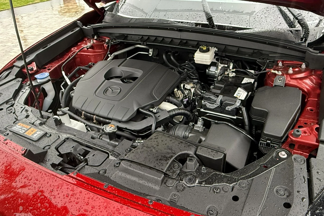 Silnik benzynowy pod maską czerwonego samochodu Mazda z wyraźnym logo na osłonie silnika, otoczenie części mechanicznych i przewodów, krople deszczu na powierzchni elementów.