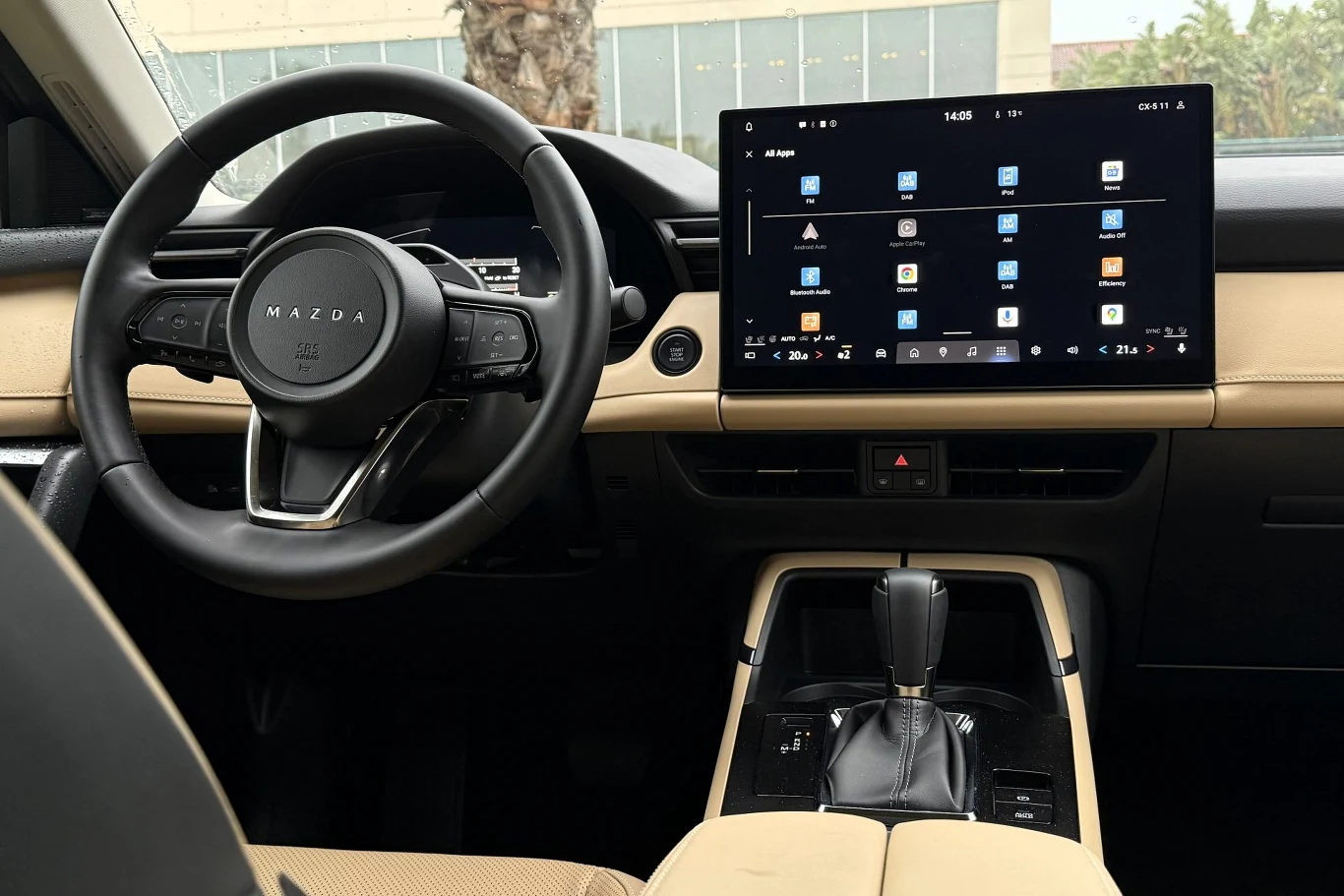 Nowoczesne wnętrze samochodu marki Mazda z dużym, dotykowym ekranem multimedialnym na konsoli centralnej, skórzaną kierownicą oraz automatyczną dźwignią zmiany biegów, utrzymane w jasnej kolorystyce z czarnymi akcentami.