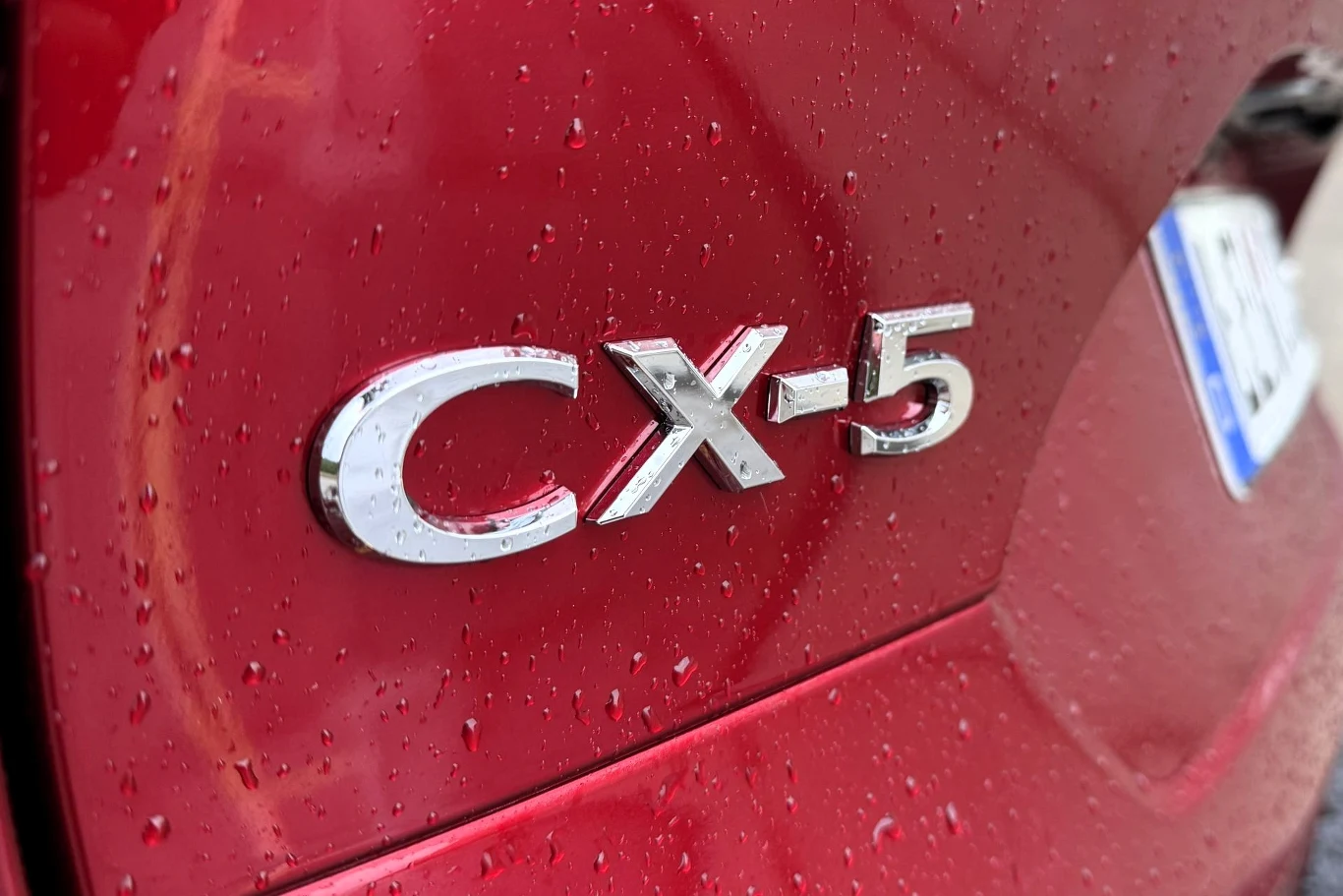 Chromowany emblemat CX-5 na czerwonym lakierze samochodu, pokrytym kroplami deszczu, obok fragment tablicy rejestracyjnej.